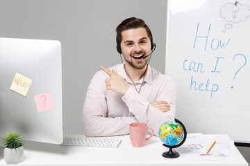 Employee operator fun business man in set microphone headset for helpline assistance sit work at call center office desk with pc computer point finger aside on area isolated on grey background studio.