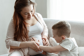 A young pregnant woman is sitting on the couch with her little son.
