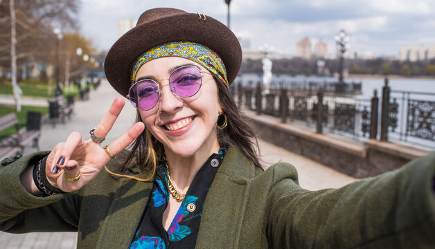 Portrait of a hippie girl in a hat and glasses who takes a selfie on a smartphone and shows the gesture of peace