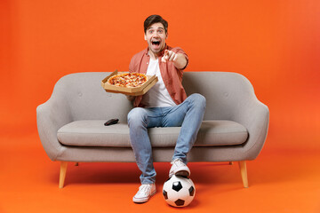 Young man football fan in shirt support team with soccer ball sit on sofa at home watch tv live...