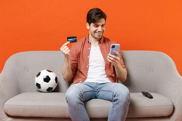 Young man football fan wearing shirt support team with soccer ball sit on sofa home watching tv live stream hold mobile cell phone credit bank card isolated on orange background. People sport concept.