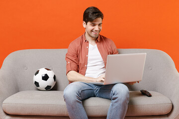Young fun man football fan wearing shirt support team with soccer ball sitting on sofa home...