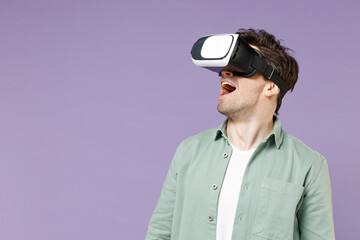 Young happy fun overjoyed man 20s wearing casual green mint shirt white t-shirt playing game watching in vr headset pc gadget isolated on purple color background studio portrait. Lifestyle concept