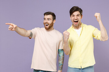Two excited amazed overjoyed young men 20s friends together wearing casual t-shirt show point index finger aside do winner gesture clench fist isolated on purple background. People lifestyle concept