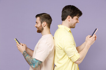Side view smiling happy young cool men friends together wearing casual t-shirt hold mobile cell phone chat online stand back to back isolated on purple background People lifestyle friendship concept.