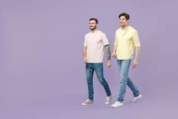 Full length side view two young happy men 20s friends together wearing casual t-shirt walking going strolling isolated on purple color background studio portrait People friendship lifestyle concept.
