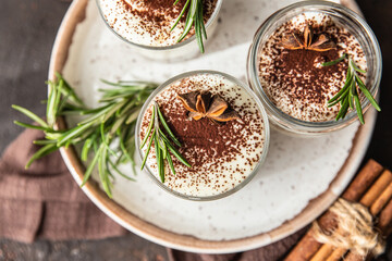 Layered dessert in glass jar with cookie crumble and whipped cream decorated with rosemary and anise, dark brown background. No bake cheesecake, trifle or pudding. Top view.