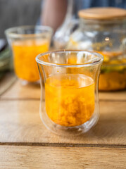 Glass cup with sea buckthorn tea and teapot on wooden table in cafe. Traditional autumn hot drink.