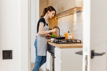 Young woman in apron cooking healthy food at modern home kitchen. Preparing meal with frying pan on gas stove. Concept of domestic lifestyle, happy housewife leisure and culinary hobby