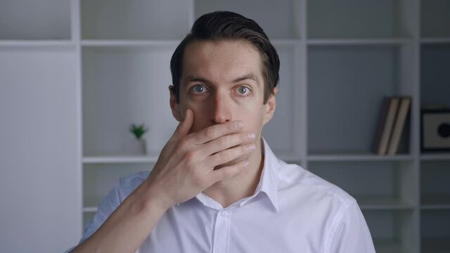 Shocked business man in white shirt takes off glasses and opening her mouth at a loss and looking to the camera on modern interior. Portrait of frightened male on office background