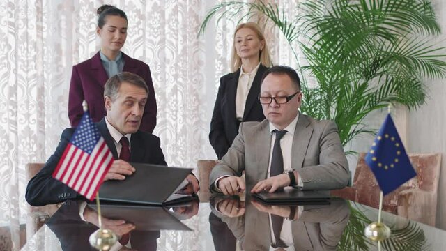 Medium slowmo of two US and UE foreign affairs ministers sitting at negotiation table, signing cooperation agreement and shaking hands while their female assistants standing behind controlling process