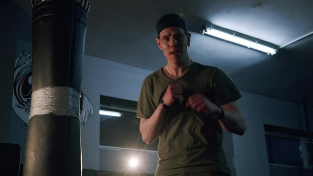 Alone boxer improves boxing movements in dark gym in slow motion. Young barefoot man training indoors on sport mats. Strong athlete in gym. Sport concept.