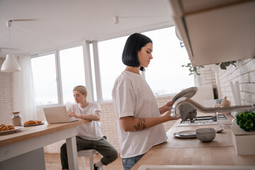 brunette woman washing plate near girlfriend using laptop on blurred background