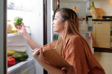 Pretty young woman checking vegetables on shelves in her fridge when making shopping list - obrazy, fototapety, plakaty