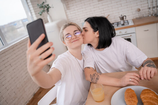 brunette woman kissing cheek of tattooed girlfriend taking selfie in kitchen
