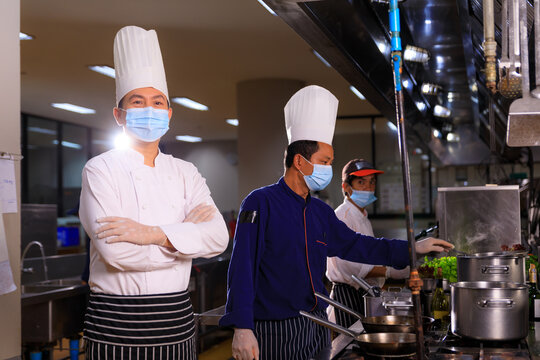 An Asian Male executive chef wearing protective mask in the kitchen with his teammate.
