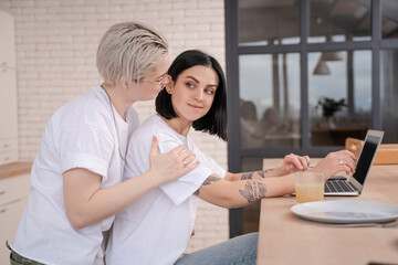 young woman looking at tattooed girlfriend using laptop in kitchen