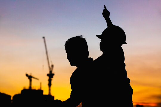silhouette of the father and son wearing a helmet Happy carrying his son on the shoulder to look at the multi storey building at sunset.