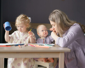 Young loving mother kissing hand of her son cute baby boy with blue eyes and golden hair while drawing with children older sister and small brother at home, family mom and kids performing art activity