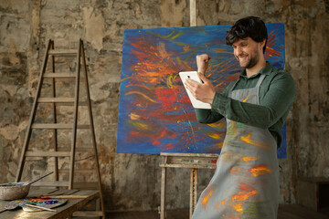 Male artist standing in his studio using a tablet wins. Workshop with oil painting