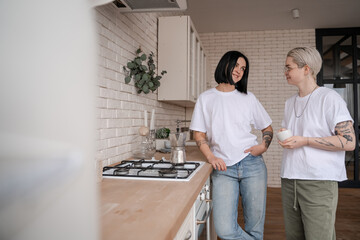 young woman holding cup and looking at girlfriend near coffee pot on stove