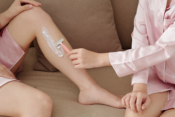 Woman using shaving cream when shaving legs of her friend with disposable razor