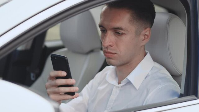 Close-up portrait of smiling male businessman sitting at the wheel of a car and using smartphone.