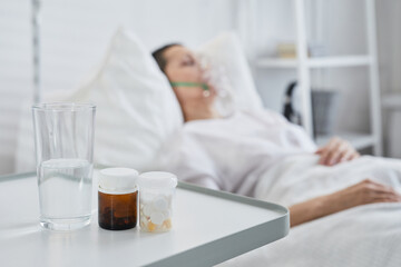 Close-up of medicines and glass of water on the table with sick patient in the background
