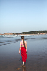 Young woman wearing a red towel and a pink bikini walking at the water edge on the beach