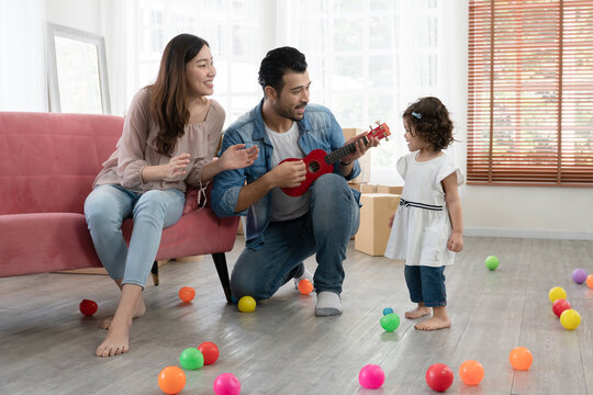 Happy Young Parents Caucasian Father And Asian Mother Playing Ukulele And Singing With Little Kid Girl In Living Room At New House. Full Of Boxes And Colorful Balls On Floor.