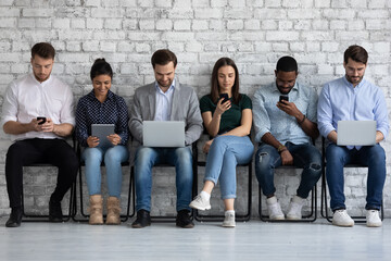 Diverse group of millennial people focused on different devices, sitting together in row, using phones and computers, checking social media news, chatting online in silence. Gadget addiction concept