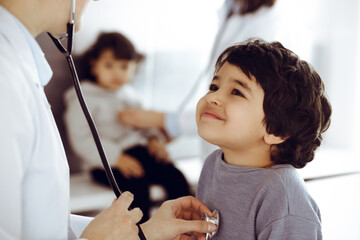 Doctor-woman examining a child patient by stethoscope. Cute arab boy and his brother at physician...