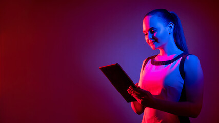 Digital communication. Future technology. Cyber lifestyle. Neon woman using tablet for teleconference in pink blue fluorescent light isolated on purple red copy space background.