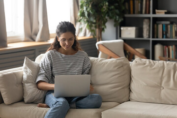 Happy young woman relax on couch at home browse wireless internet on laptop. Smiling millennial female in glasses work online on computer, talk speak on video call. Technology concept.