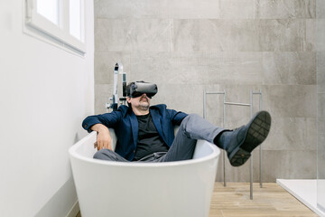 Fully clothed elegant adult male experiencing virtual reality in a bathtub as if he were on a tropical beach. Virtual reality glasses. Virtual reality holiday.