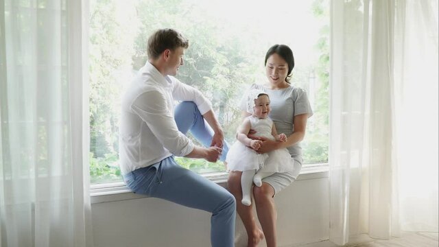 Mother, father, and baby posing for the family photo in studio, holding a baby on lap. Mixed race family posing for the family picture while sitting in festive clothes on the windowsill, daytime