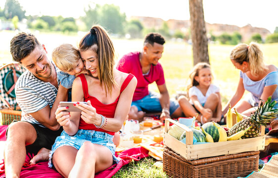 Happy multiethnic families playing with phone at pic nic garden party - Joy and love life style concept with mixed race people having fun together at picnic barbecue before sunset