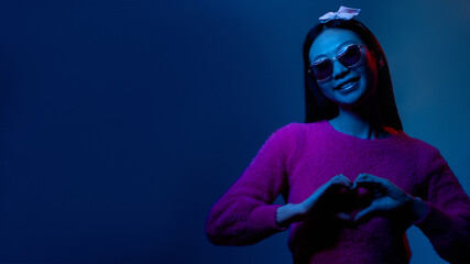 Color light portrait. Love sign. Romantic sympathy. Blue neon supportive grateful smiling Asian fan girl in pink showing heart gesture isolated on dark night copy space background.