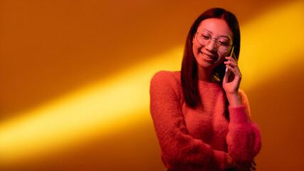 Phone call. Color light banner. Mobile communication. Neon pink smiling Asian woman in glasses enjoying conversation isolated on yellow golden gradient copy space background.