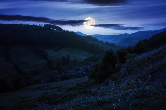rural valley landscape at night. beautiful carpathian nature scenery with grassy hills, fields and meadows between forested hills in full moon light. small village in the distance