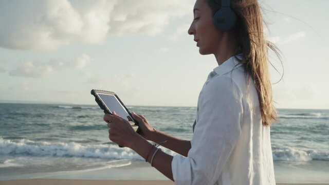 Tracking medium shot of brown-haired Caucasian woman wearing over-ear headset, using tablet computer for video calling, walking by sunny windy ocean shore, smiling