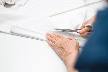 Elderly woman cutting white fabric with scissors, hands close-up. Senior woman dressmaker. Retired work or hobby at home.