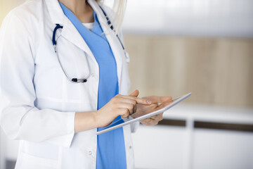 Unknown woman-doctor standing in sunny clinic and using tablet pc, close-up. Data and best service in medicine