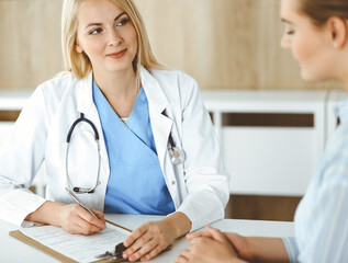Woman-doctor and patient sitting and talking in hospital or clinic. Blonde therapist is cheerfully...