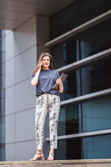 Beautiful woman with brown long hair in elegant casual clothes with a laptop speaks on a mobile phone outdoors with office buildings in the background