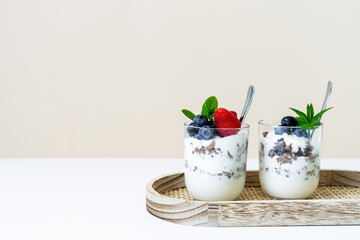 Zwei Gläser mit Joghurt, Granola und frischen Beeren geschichtet auf einem Holz Serviertablett. Weißer Tisch, beige Wand, modern, gesundes Frühstück.