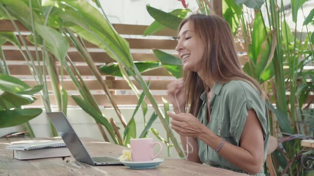 Waist-up of young Caucasian woman sitting at table in outdoor cafe, untangling wired headset, working, starting online video call on laptop computer, smiling