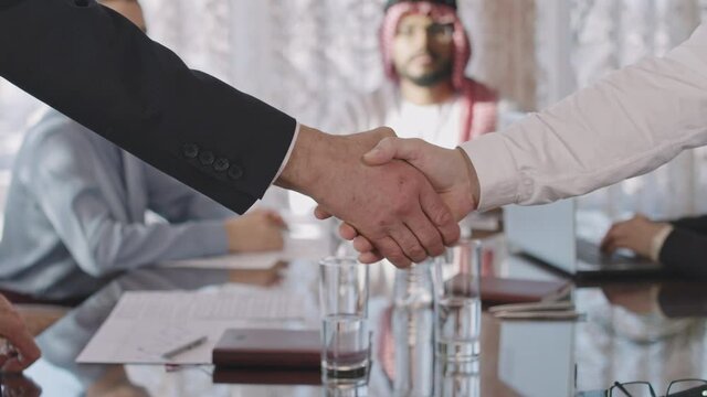 Slowmo PAN close-up of unrecognizable male investors shaking hands after making successful deal during negotiations with diverse business partners in modern conference room