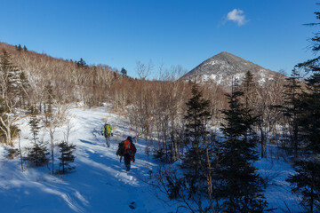 The snowy peaks of the Sestra mountain in the Lazovsky district of the Primorsky Territory. Two tourists walk along a snowy path among the trees to the top of the mountain.