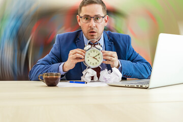 Boss businessman at his desk in the office with a bunch of crumpled paper holds an alarm clock in his hands. Strenuous office work. Preparing of report. Stress. Blurred abstract background.